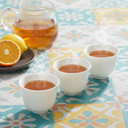 Three white handleless ceramic tea cups with a glass teapot on a colorful patterned table